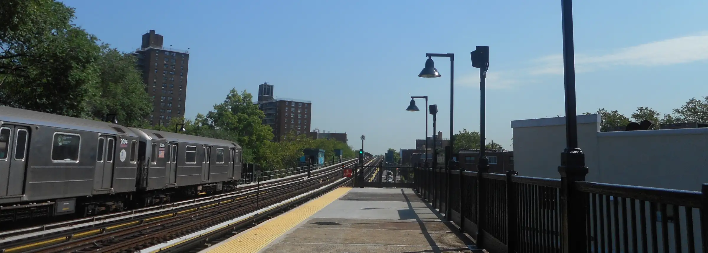 A 6 train stopped at the elevated Middletown Road station, with trees and apartment towers behind, on a sunny morning